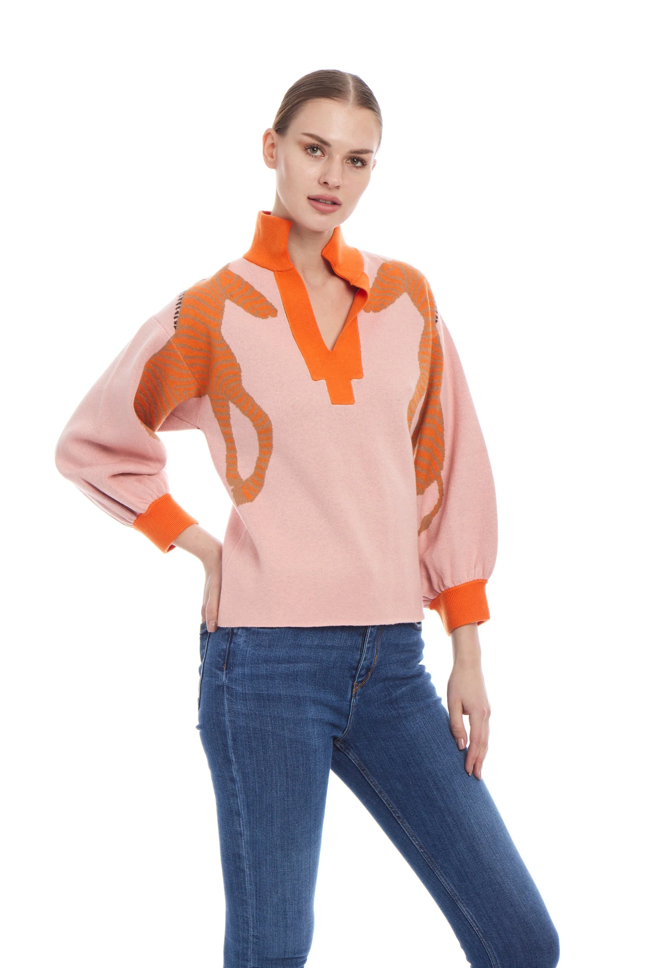 A woman modeling a light pink knit sweater with bold orange graphic patterns and a high orange collar, paired with blue jeans, against a plain white background.