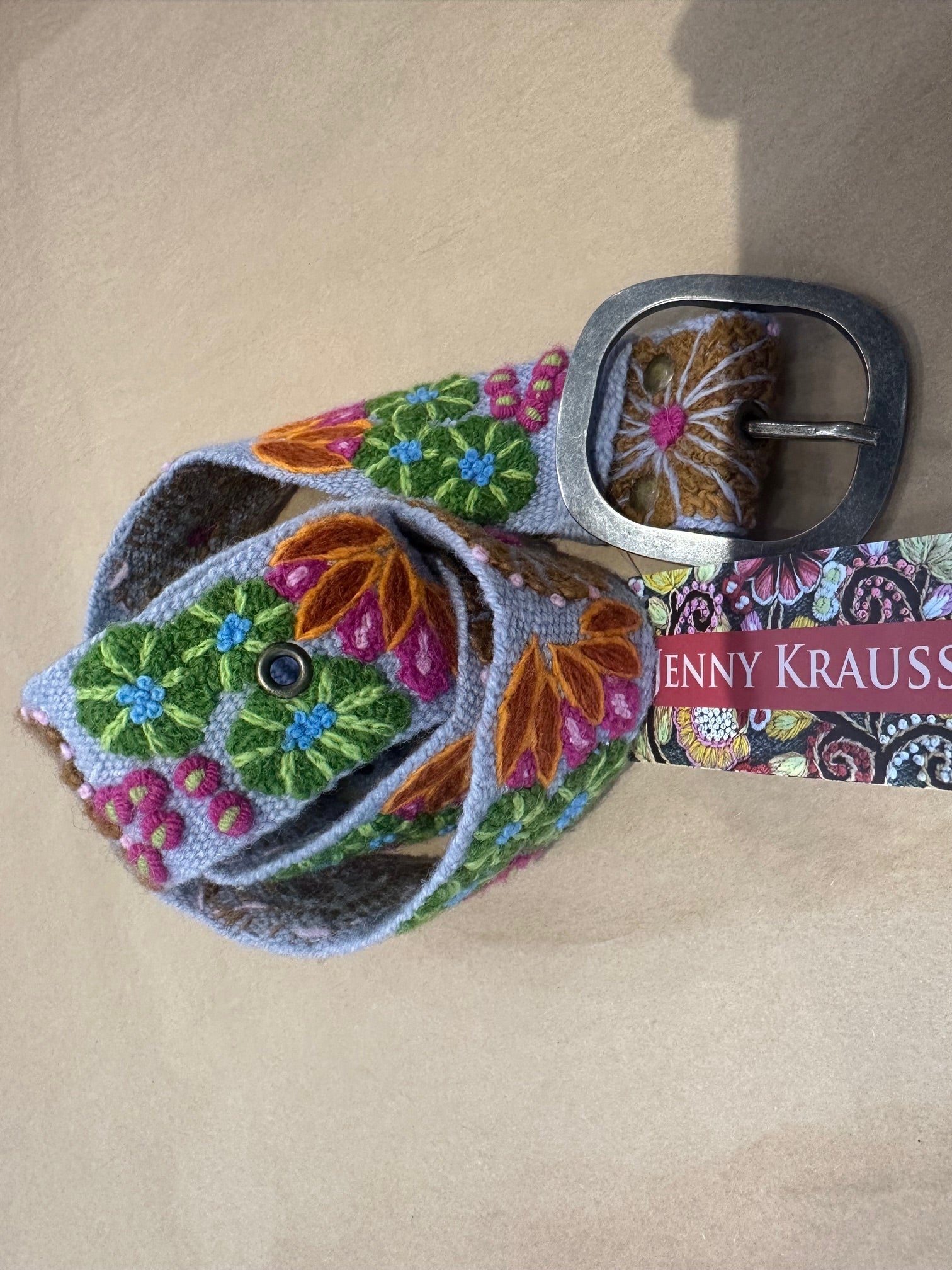 Colorful floral-patterned belt with a silver buckle on a beige surface, featuring the brand 'Jenny Krauss'.
