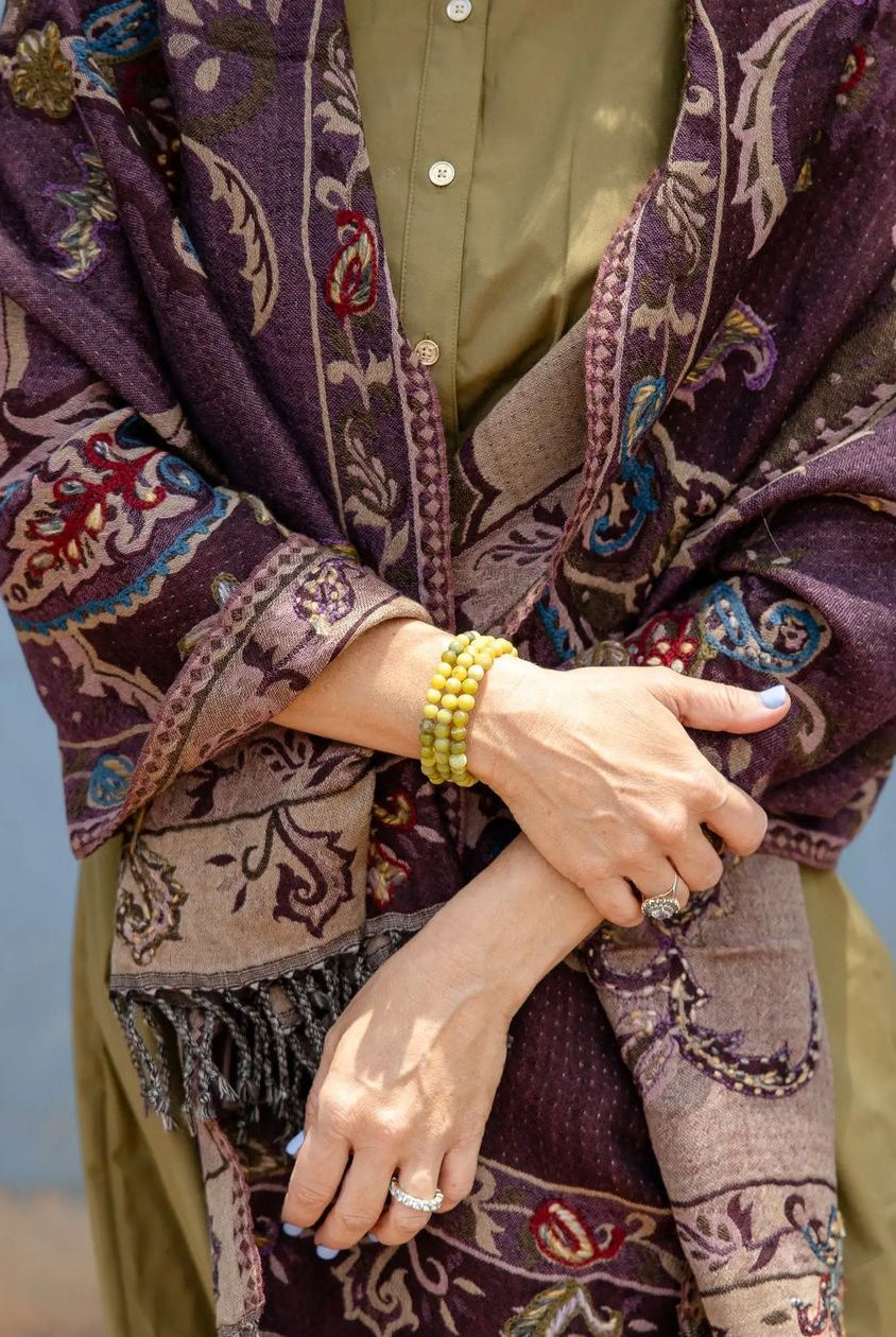 A close-up of a person wearing a patterned shawl in deep purple, beige, and brown over an olive green kurta, with yellow beaded bracelets on one wrist.
