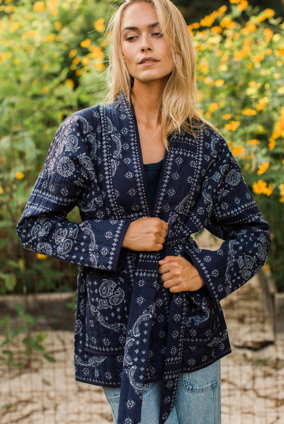 A woman wearing a navy blue long-sleeve wrap jacket with a white paisley bandana print, standing outdoors in front of a blurred background of green foliage and yellow flowers.