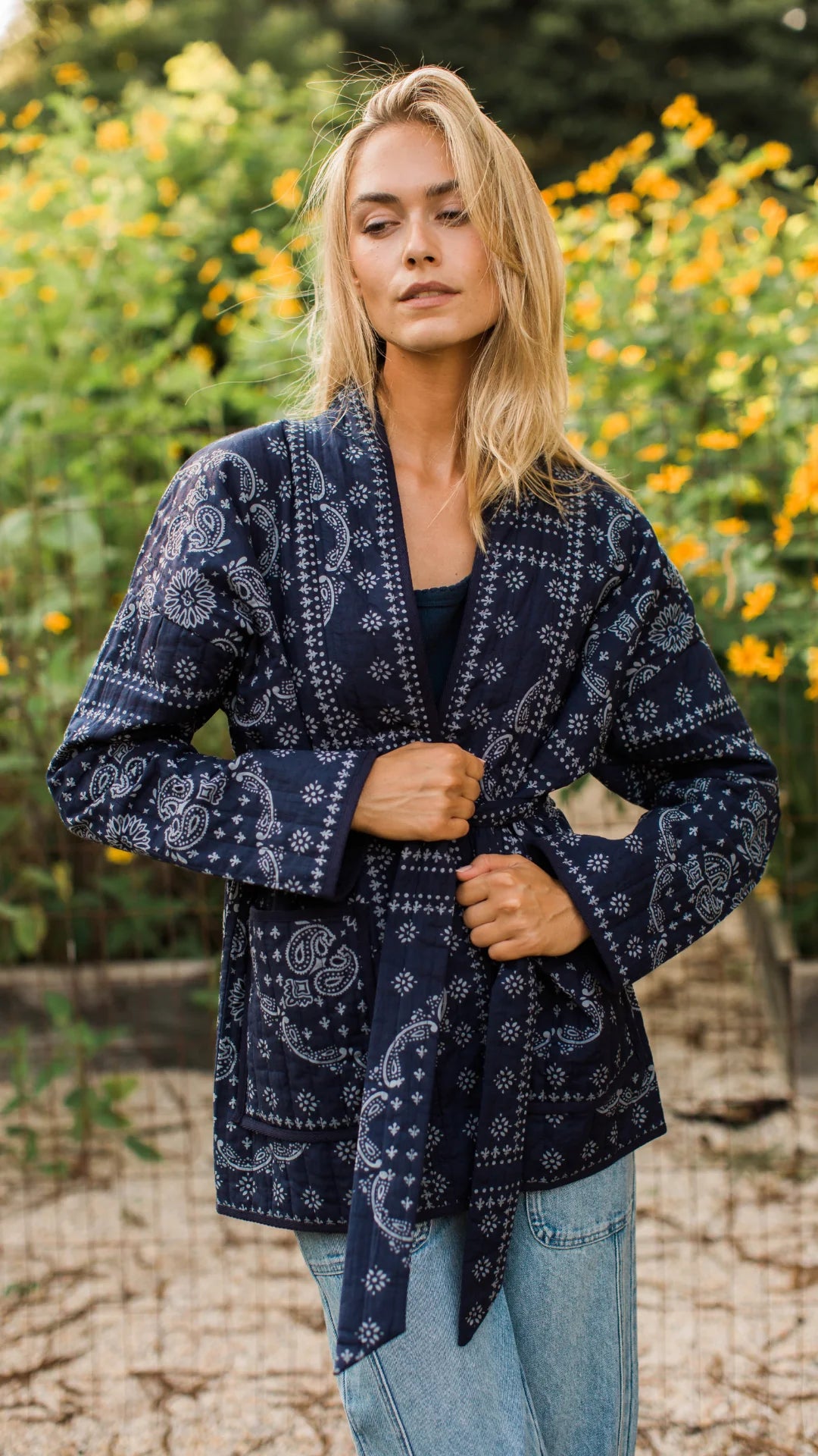 A woman wearing a navy blue long-sleeve wrap jacket with a white paisley bandana print, standing outdoors in front of a blurred background of green foliage and yellow flowers.