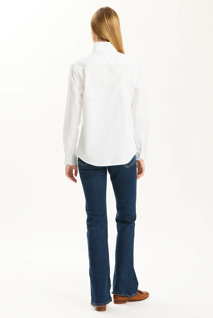 A woman viewed from the back wearing a white long-sleeve button-up shirt, dark blue flared jeans, and brown shoes against a plain white studio background.