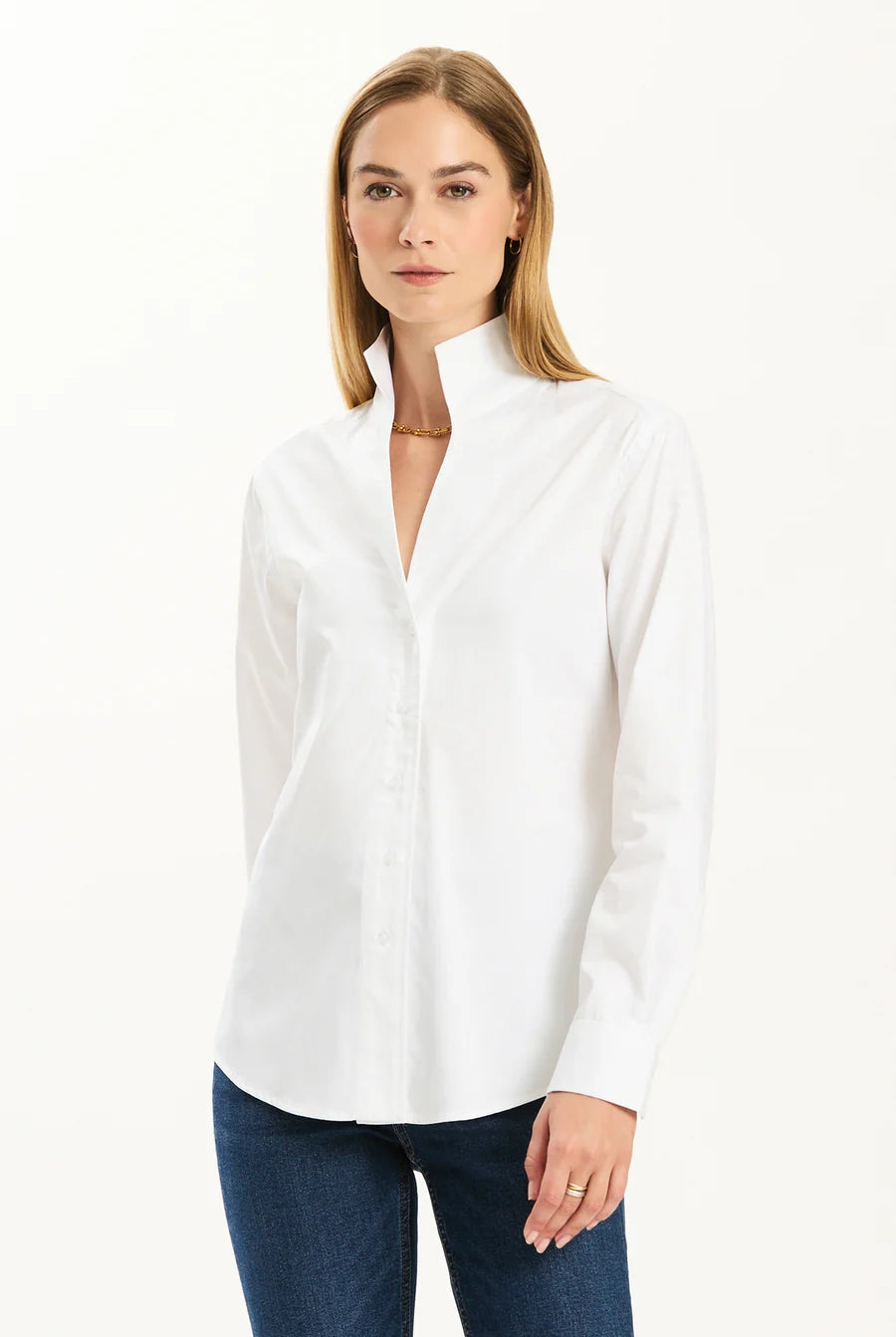A woman wearing a white long-sleeve button-up shirt with a high stand collar and dark blue jeans, standing against a plain white studio background.