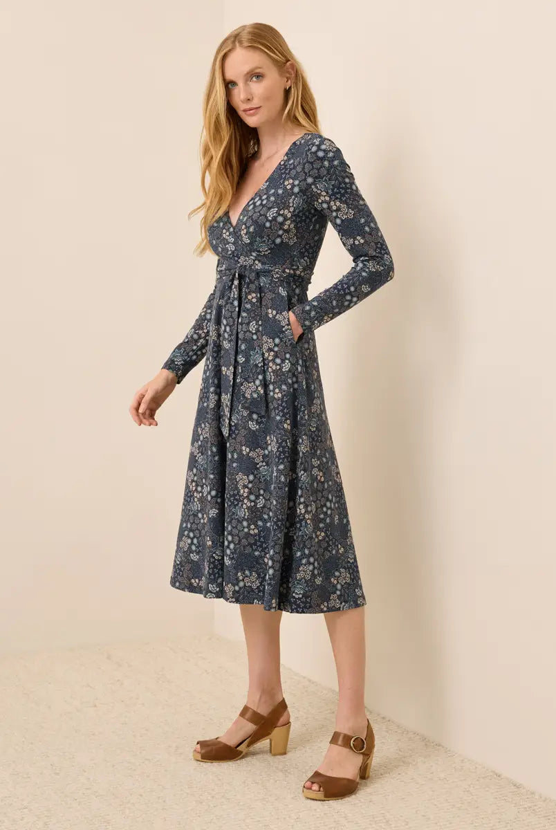 A woman modeling a long-sleeve wrap-style midi dress with a blue floral print, standing against a plain beige wall and textured floor.