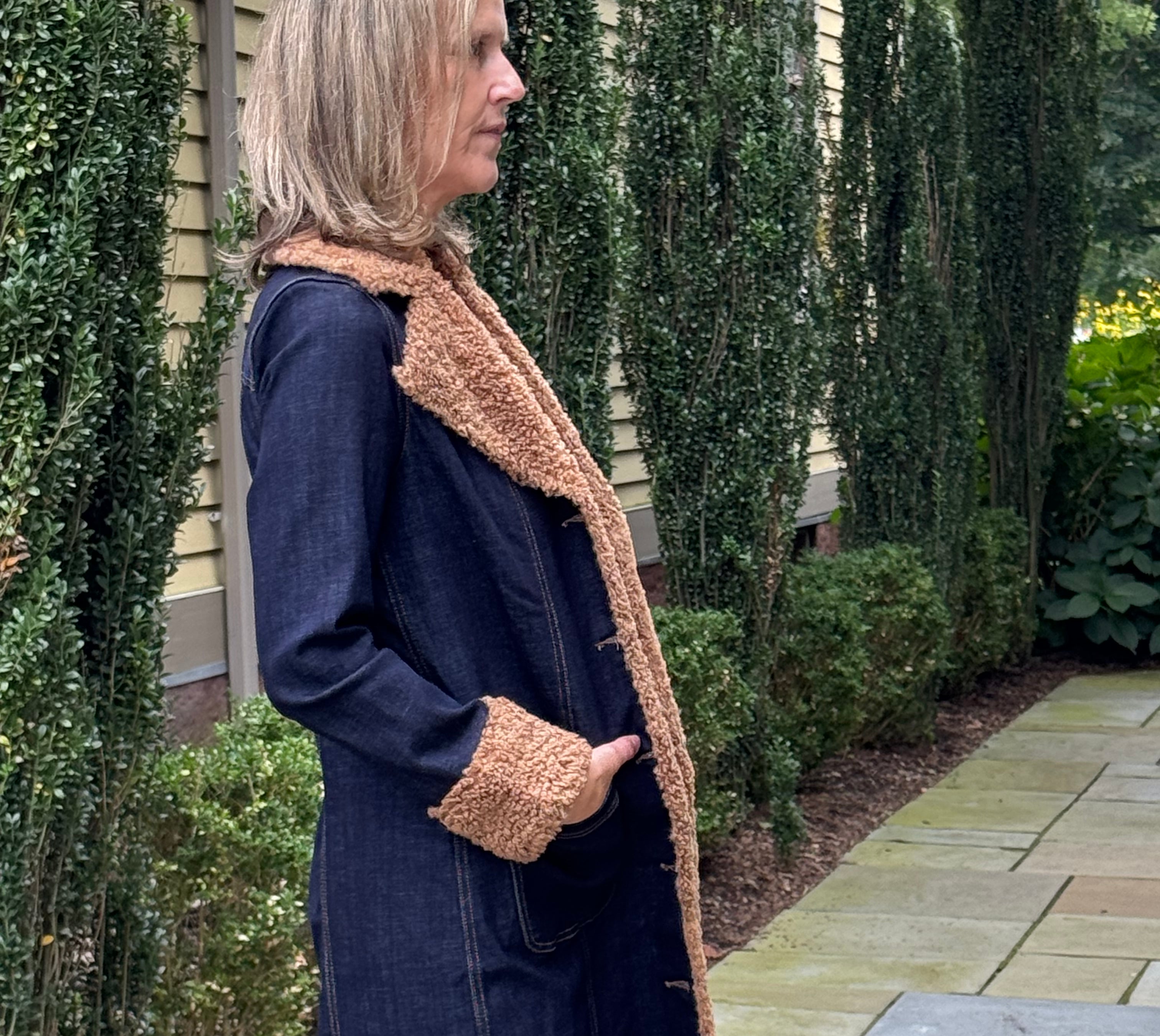 A woman wearing a dark navy blue denim coat with a tan sherpa lining, standing on a stone patio in front of a house with tall green hedges.