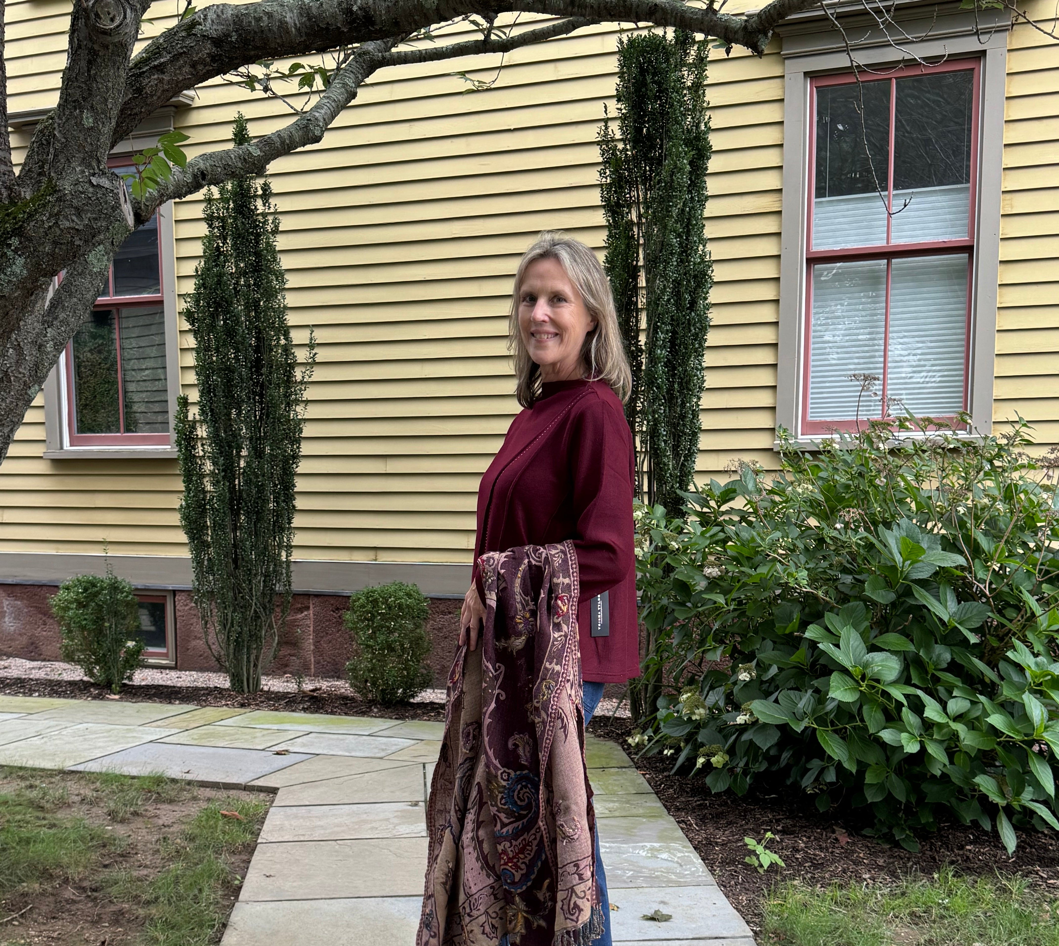 A woman stands on a stone walkway in front of a yellow house, holding up a long patterned scarf or shawl over her arm. She is wearing a burgundy top, blue jeans, and black sandals.