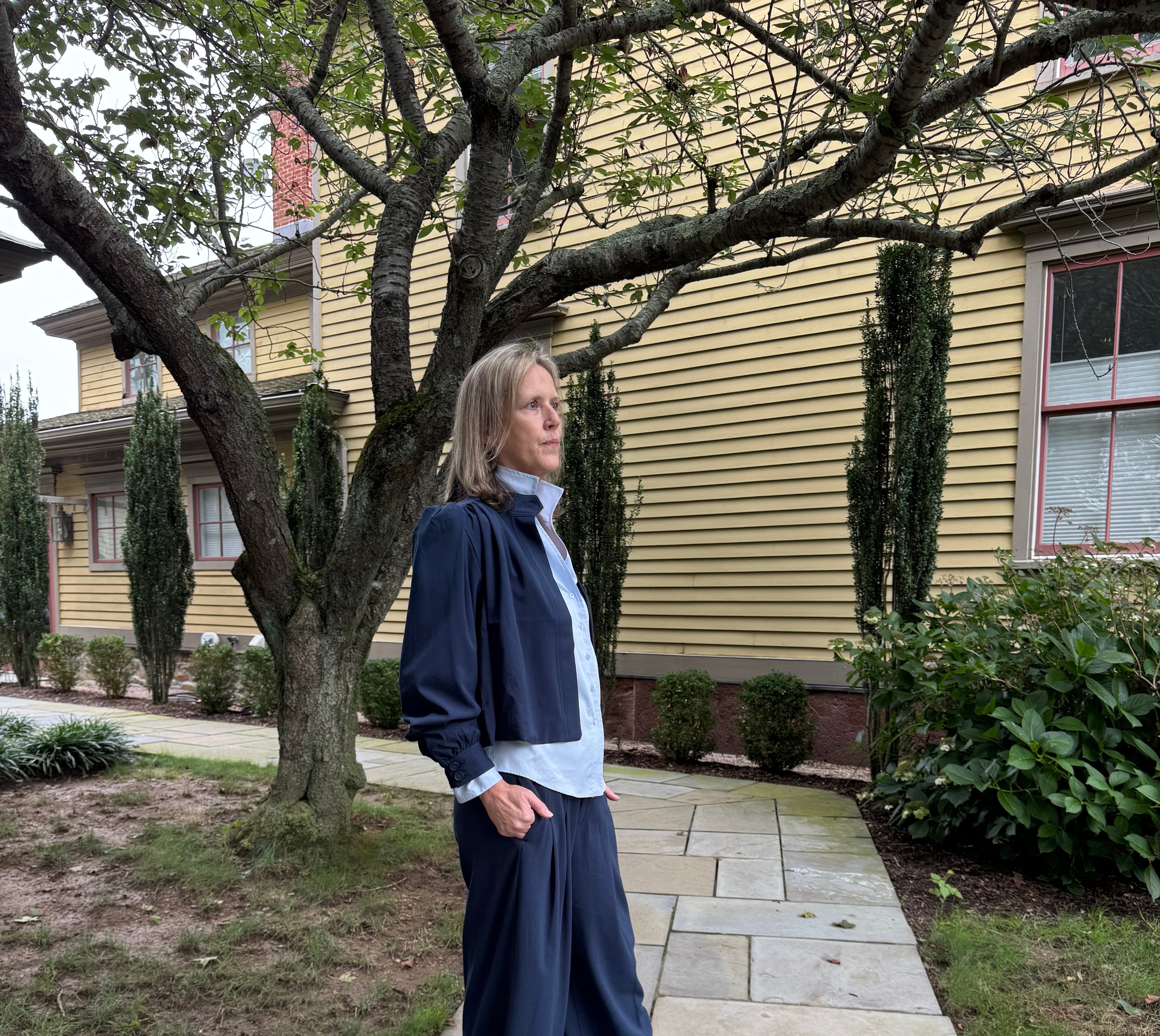 A woman stands on a stone walkway in front of a yellow house, wearing a navy blue wide-leg skirt or pants, a light blue button-up shirt, and black sandals.