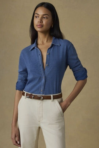 A woman modeling a blue long-sleeve button-up shirt with cream trousers and a brown belt, standing against a warm beige studio background.