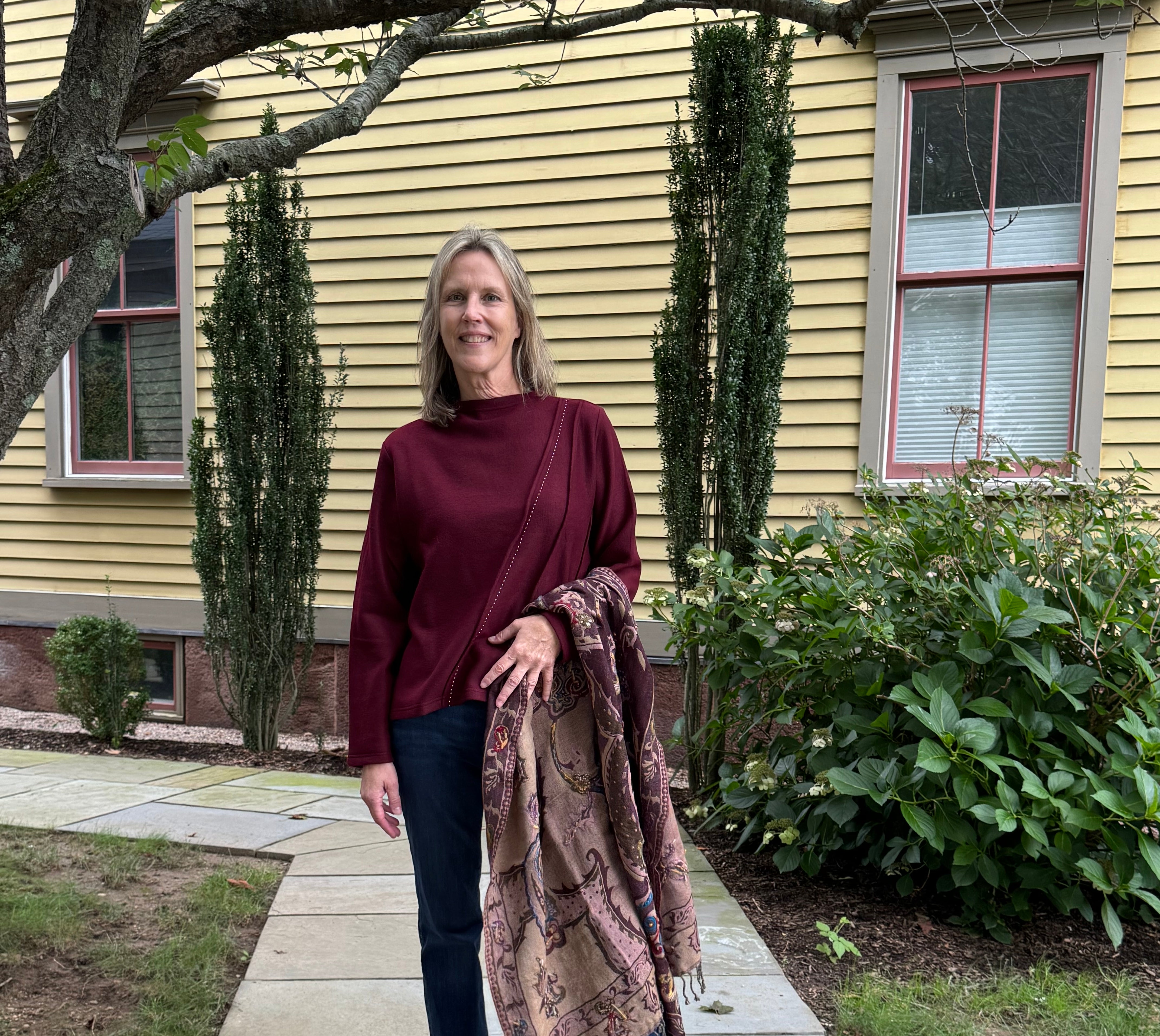 A woman stands on a stone walkway in front of a yellow house, wearing a burgundy mock-neck top, dark blue jeans, and black sandals, while holding a brown and beige patterned shawl.