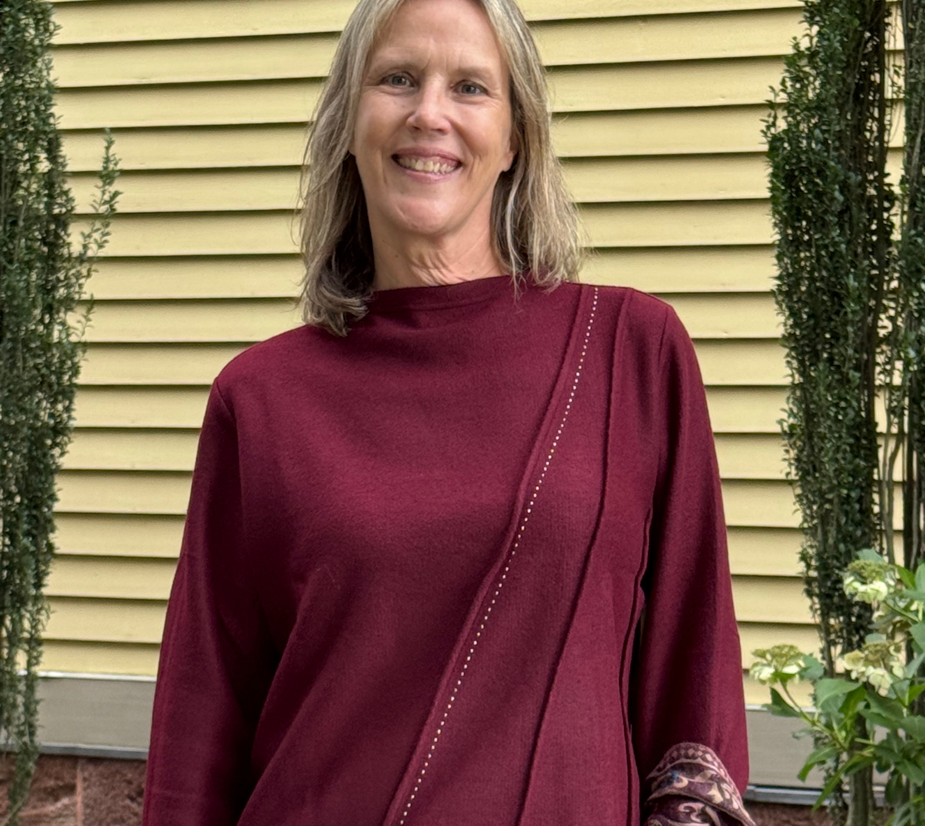 A smiling woman with blonde hair stands outdoors in front of a yellow house wall, wearing a long-sleeve mock-neck top in deep burgundy with diagonal seam detailing and blue jeans.