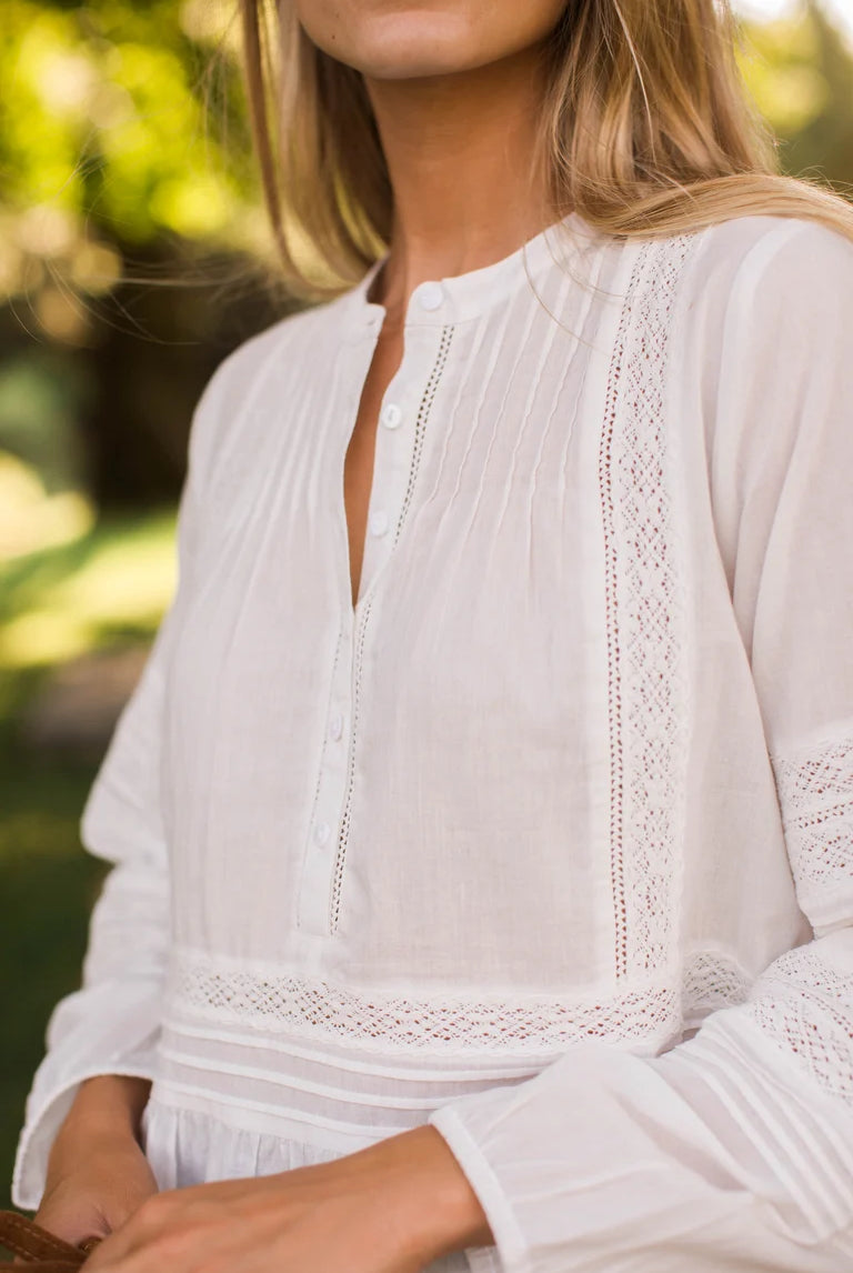 A close-up of a woman wearing a white long-sleeve blouse with delicate eyelet lace trim and a split neckline, photographed outdoors against a softly blurred green background.