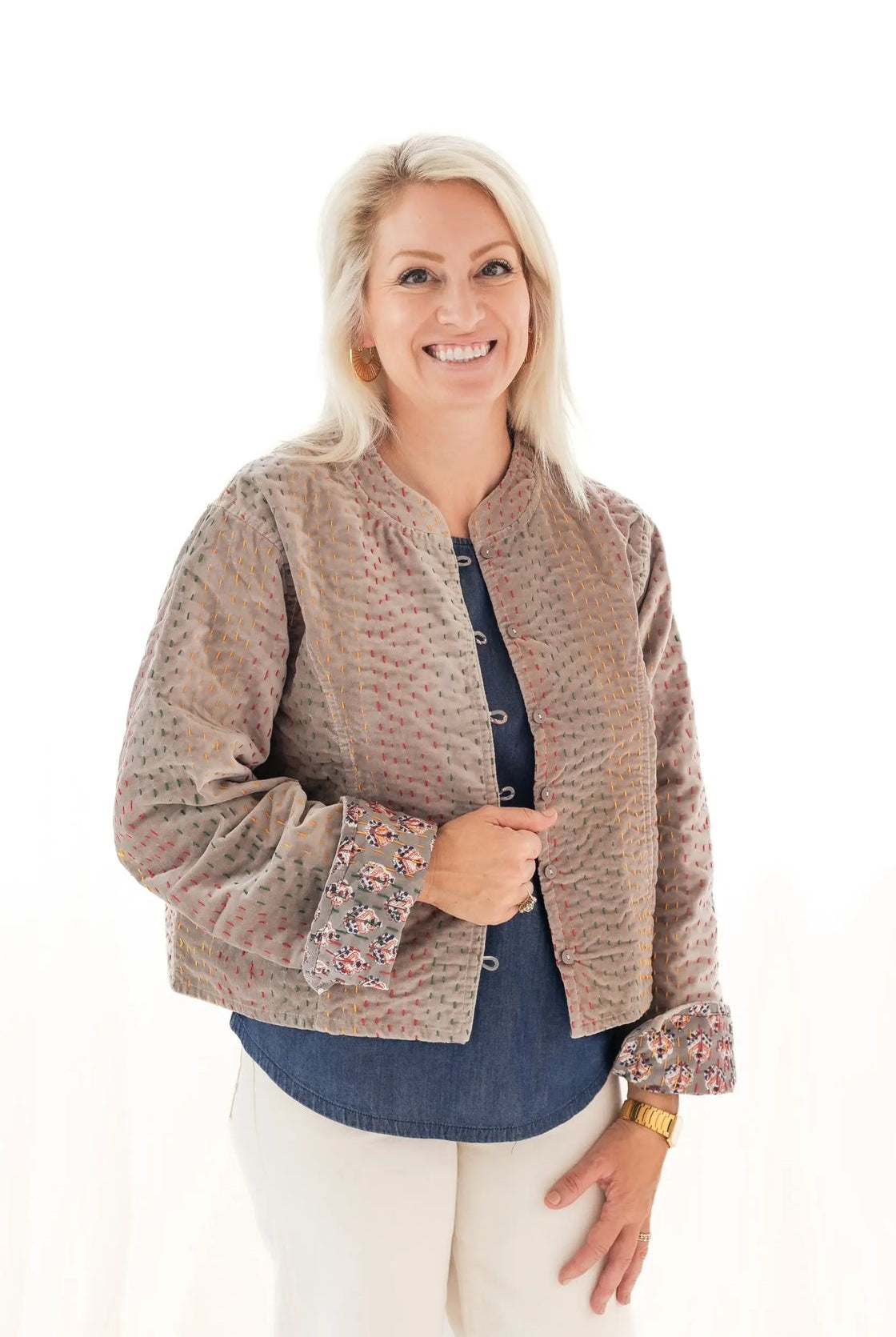 A smiling blonde woman wearing a cropped, textured beige jacket with floral-patterned cuffs over a blue button-up shirt and cream pants, photographed against a plain white background.