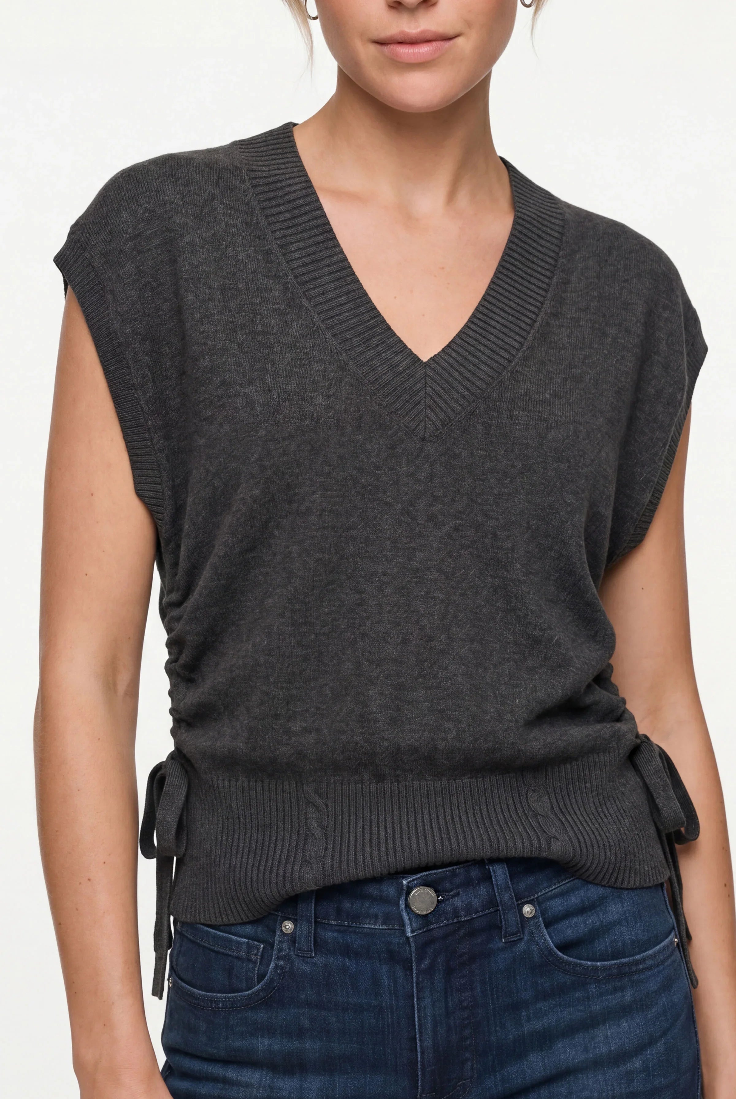 A woman wearing a dark charcoal gray sleeveless V-neck knit sweater vest with side tie details, paired with dark blue jeans, against a plain light background.