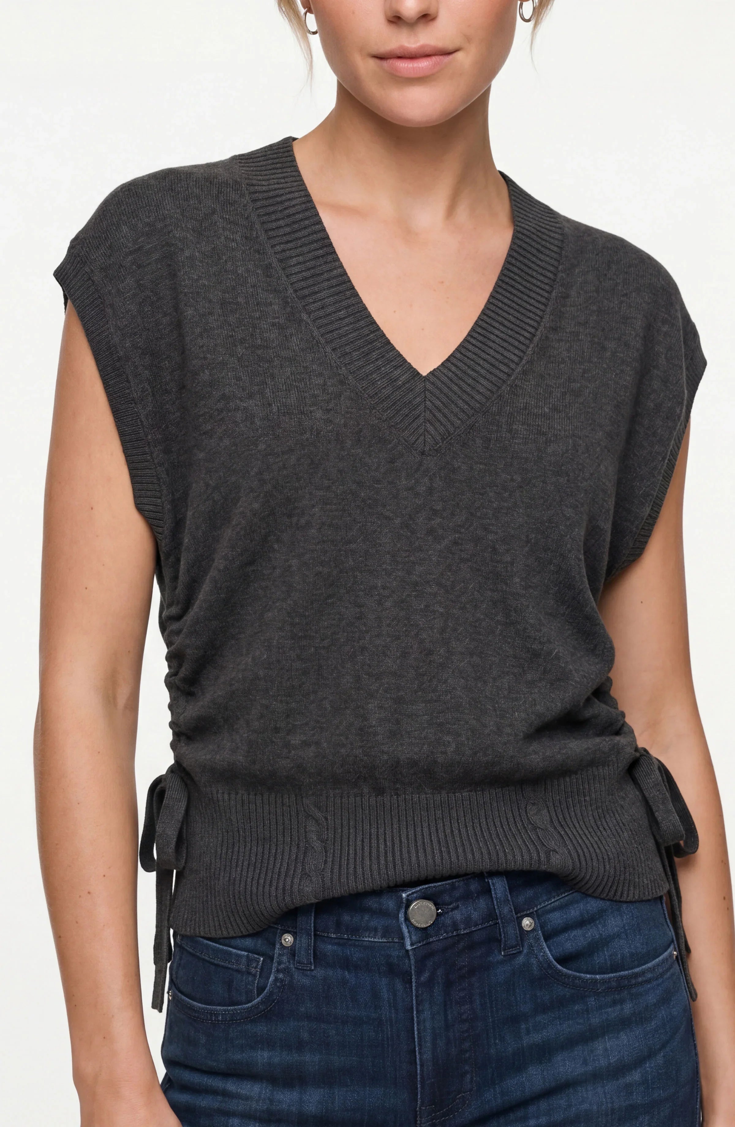 A woman wearing a dark charcoal gray sleeveless V-neck knit sweater vest with side tie details, paired with dark blue jeans, against a plain light background.