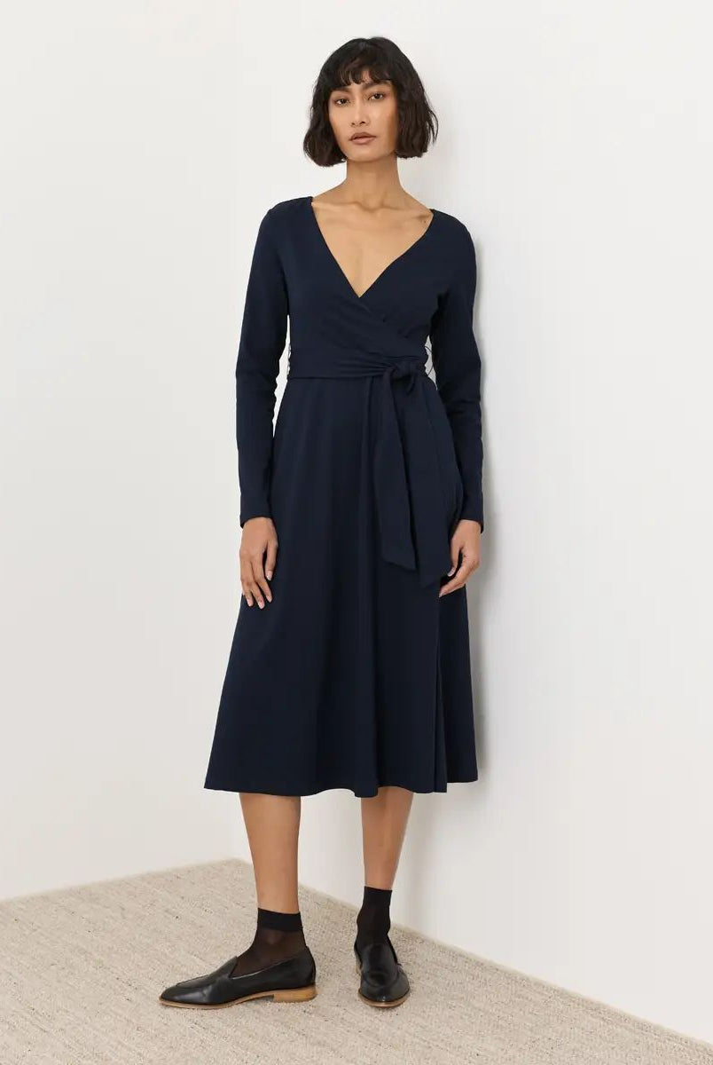 A woman modeling a dark navy long-sleeve wrap midi dress with a tie waist, paired with black socks and leather shoes, standing against a plain white wall.