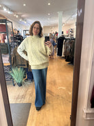 A woman takes a mirror selfie in a clothing boutique, wearing a cream cable-knit mock-neck sweater and wide-leg blue jeans.