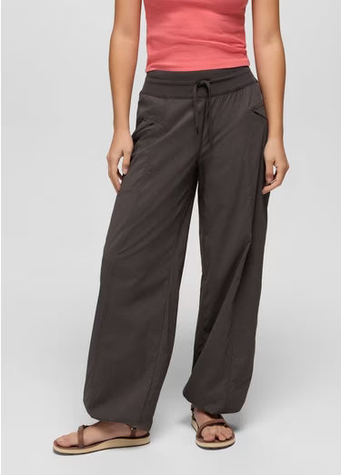 A model wearing dark brown wide-leg drawstring pants, a coral pink tank top, and brown sandals, standing against a light gray studio background.
