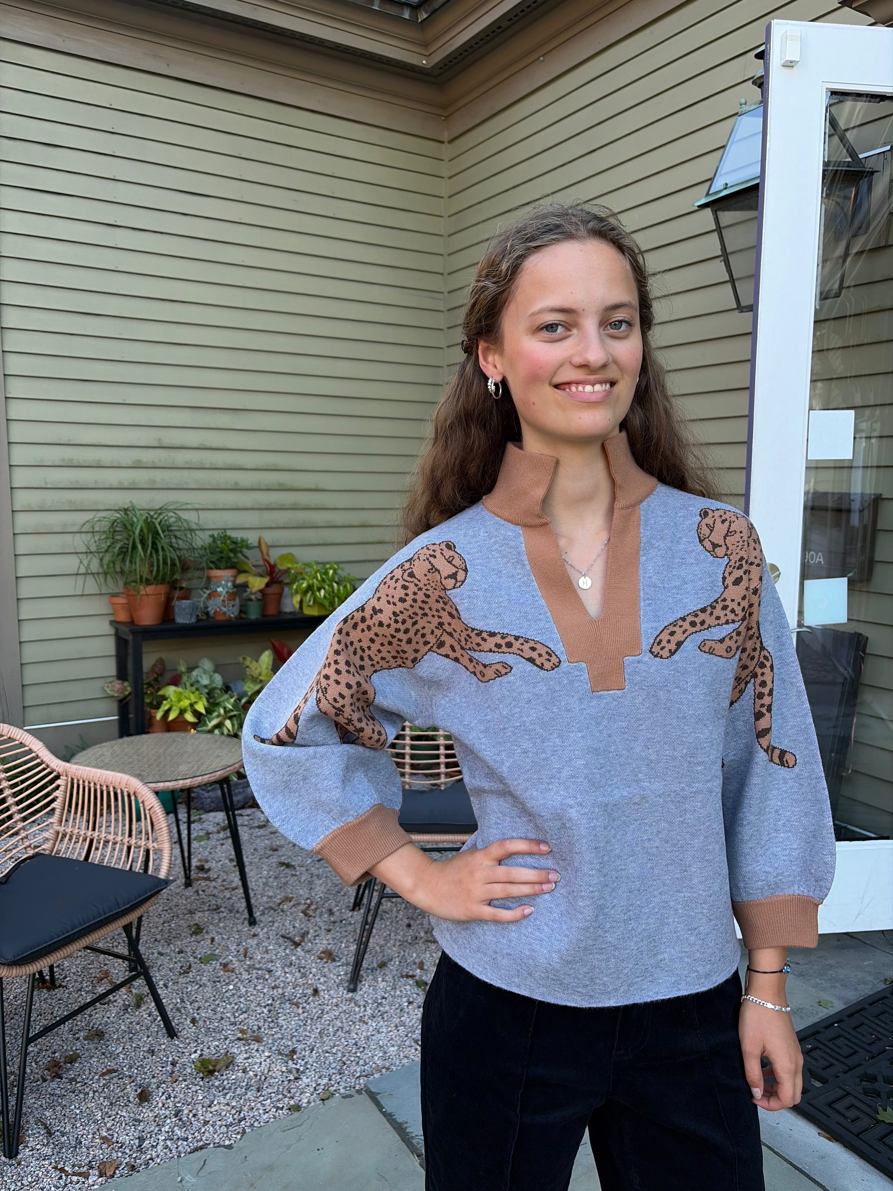 A woman wearing a light blue knit sweater with brown cheetah prints on the shoulders and brown ribbed trim, standing on a patio in front of a house with beige siding.