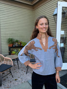 A woman wearing a light blue knit sweater with brown cheetah prints on the shoulders and brown ribbed trim, standing on a patio in front of a house with beige siding.