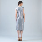 A woman stands with her back to the camera wearing a sleeveless, knee-length silver dress with a subtle speckled texture, paired with beige high heels, against a light gray studio background.