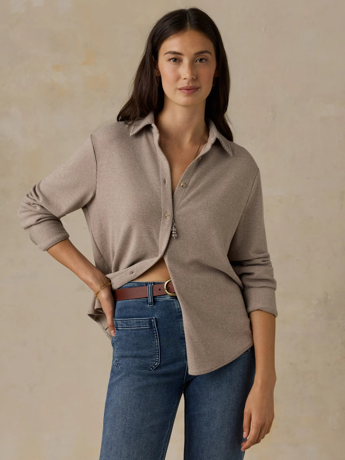 A woman modeling a light taupe button-up knit top with rolled sleeves and a relaxed fit, paired with blue jeans and a brown belt, against a textured beige studio backdrop.