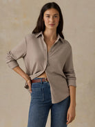 A woman modeling a light taupe button-up knit top with rolled sleeves and a relaxed fit, paired with blue jeans and a brown belt, against a textured beige studio backdrop.