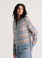 A woman wearing a light blue knit zip-up cardigan with pink and white geometric patterns, paired with light-wash blue jeans, against a plain light gray background.