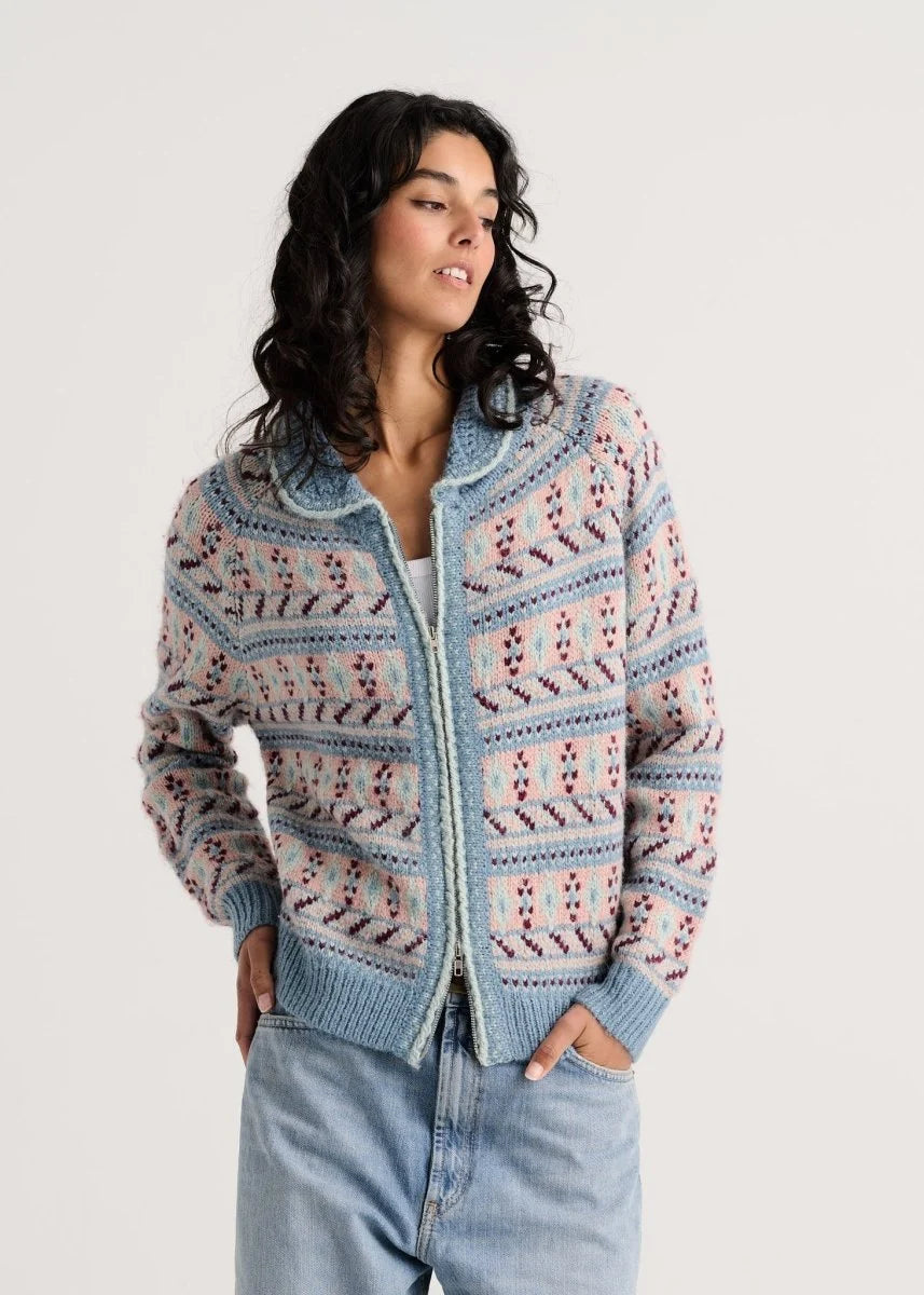 A woman wearing a light blue knit zip-up cardigan with a pink and dark blue geometric pattern, paired with light-wash blue jeans, against a plain light gray background.