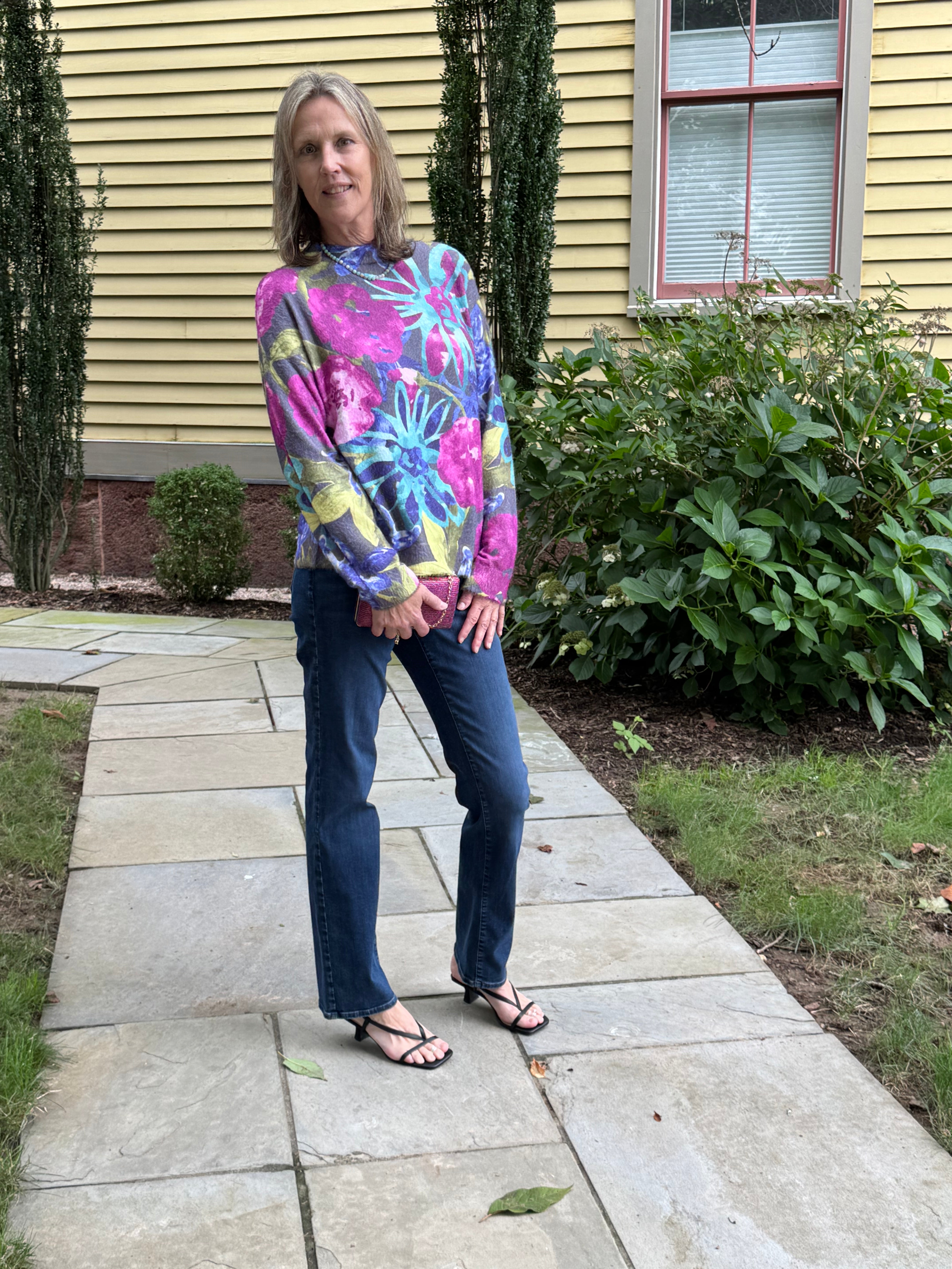 A woman stands on a stone walkway in front of a yellow house, wearing a colorful long-sleeve floral sweatshirt, blue jeans, and black sandals.