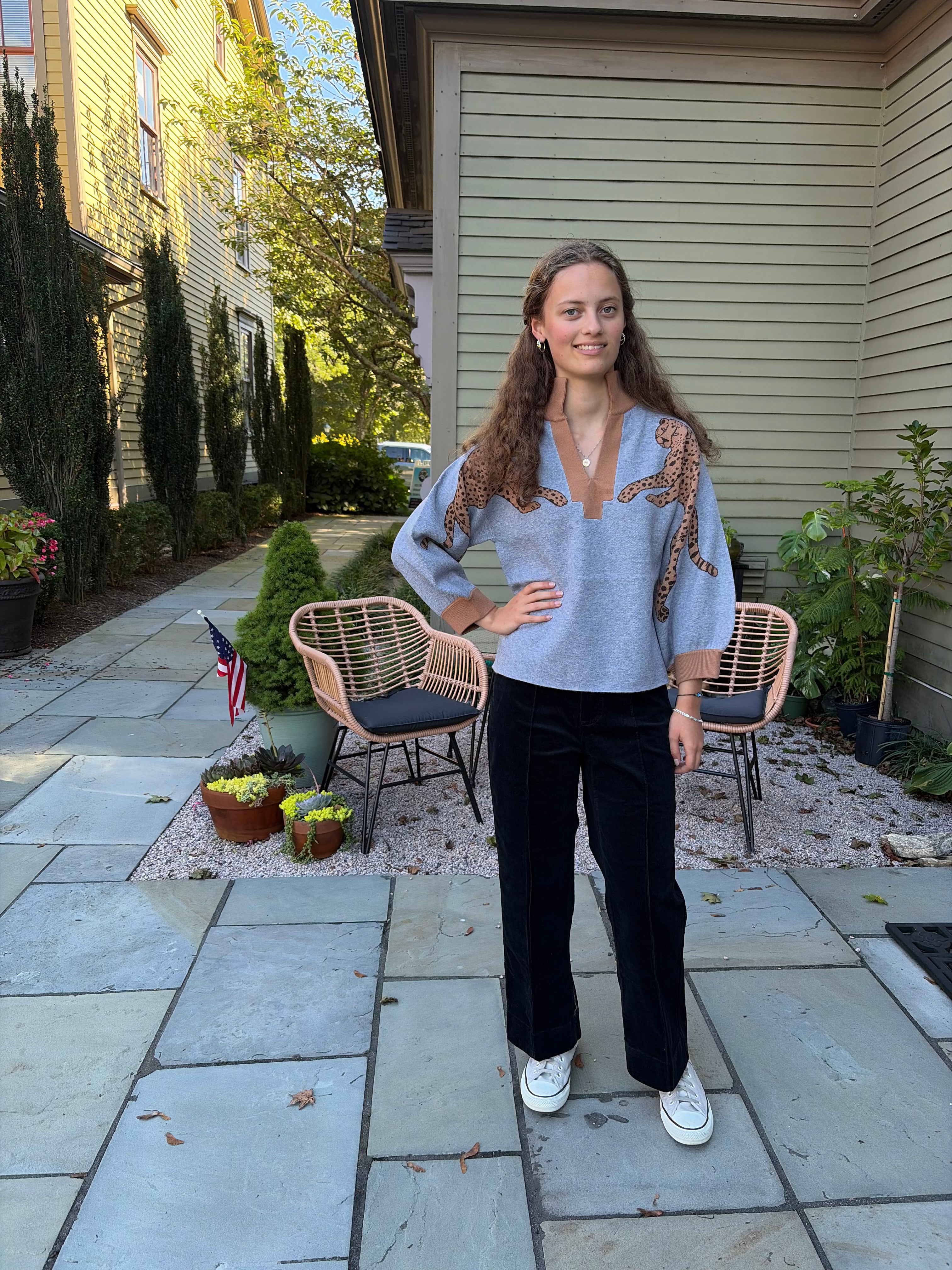 A woman stands on a stone patio in front of a house, wearing a light blue knit sweater with brown and gold patterned shoulder details, black wide-leg pants, and white sneakers.