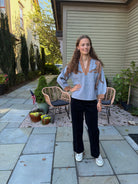 A woman stands on a stone patio in front of a house, wearing a light blue knit sweater with brown and gold patterned shoulder details, black wide-leg pants, and white sneakers.
