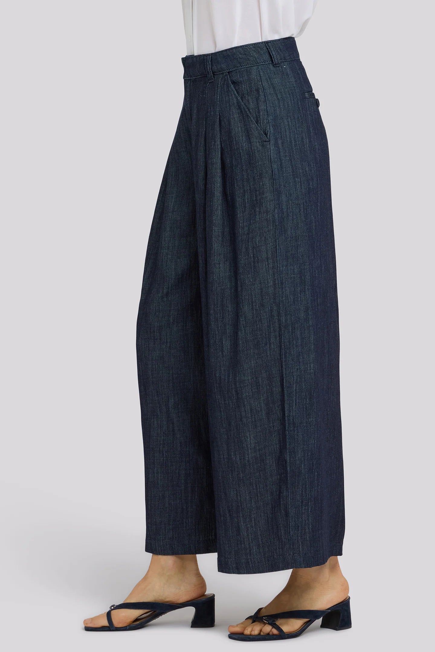 A person wearing dark blue wide-leg cropped pants with pleats, styled with a white top and black mule sandals against a light gray studio background.
