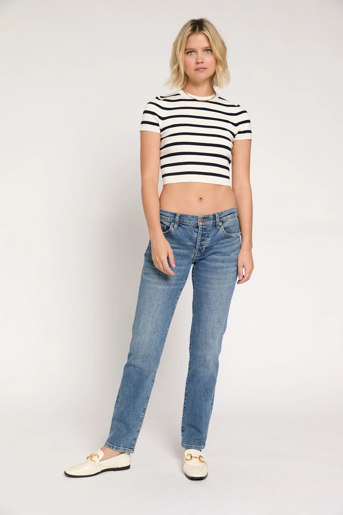 A woman modeling a black and white horizontally striped short-sleeve crop top with blue jeans and cream-colored loafers, standing against a plain light gray studio background.