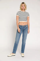 A woman modeling a black and white horizontally striped short-sleeve crop top with blue jeans and cream-colored loafers, standing against a plain light gray studio background.
