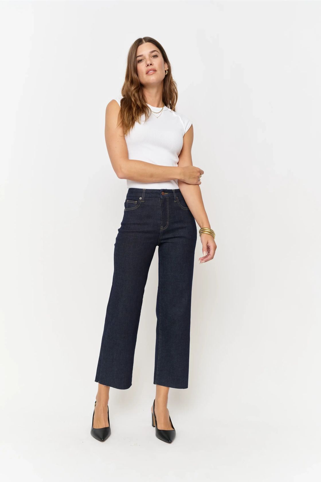 A woman modeling a pair of dark wash cropped wide-leg jeans, paired with a white sleeveless top and black heels, against a plain white studio background.
