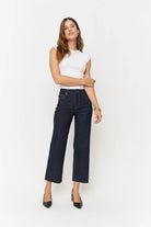 A woman modeling a pair of dark wash cropped wide-leg jeans, paired with a white sleeveless top and black heels, against a plain white studio background.