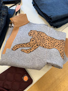 A folded heather gray sweatshirt with a large tan and black cheetah graphic across the chest, tan ribbed collar and cuffs, and a light wood-toned surface underneath. Other folded garments are partially visible around it.