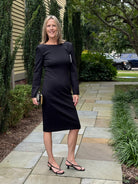 A woman stands on a stone walkway outdoors wearing a fitted black long-sleeve knee-length dress with structured shoulders, paired with black strappy heels and holding a small metallic clutch.