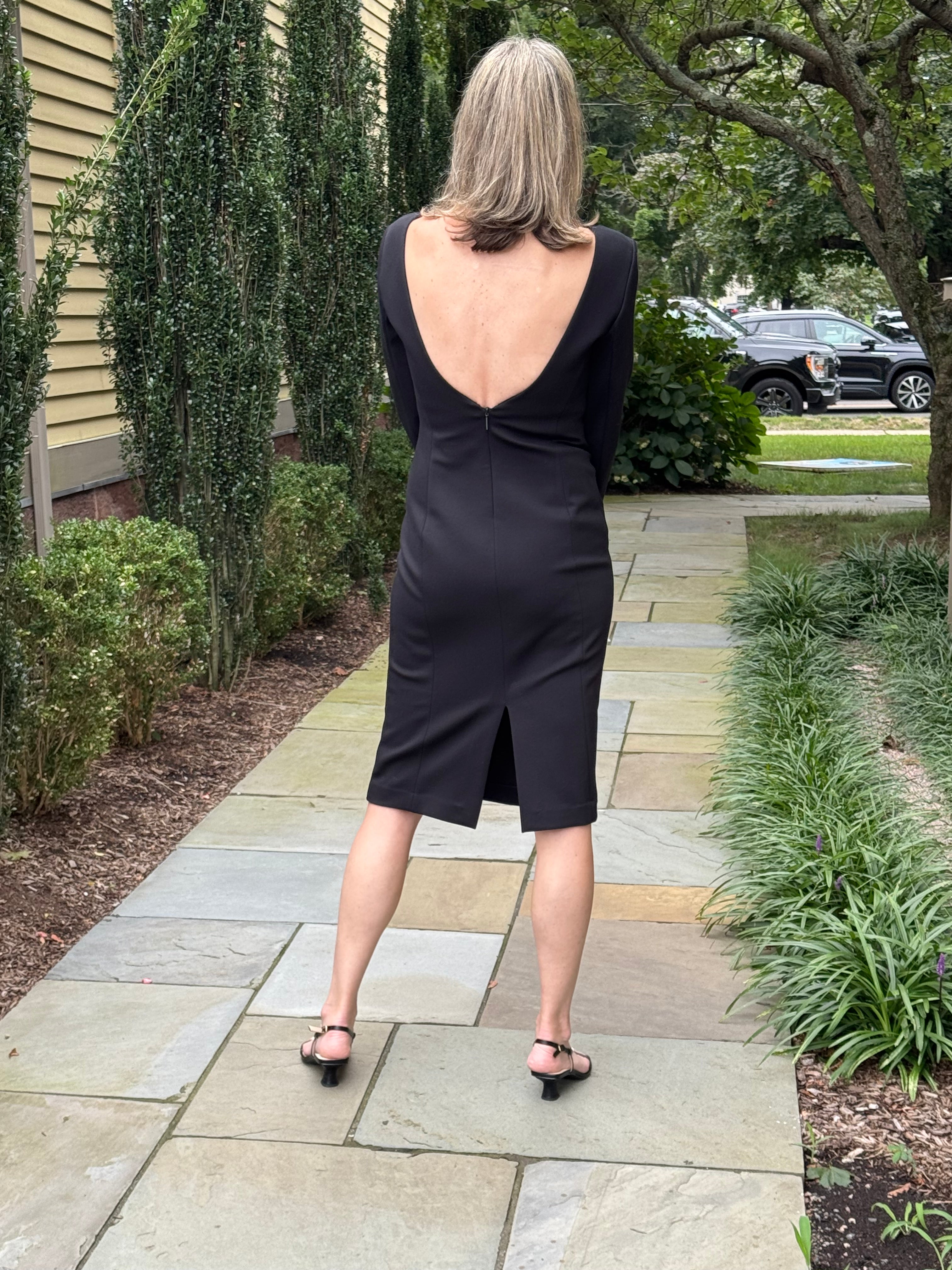 A woman stands on a stone walkway outdoors, photographed from behind, wearing a black long-sleeve dress with a deep open back and a small front slit.