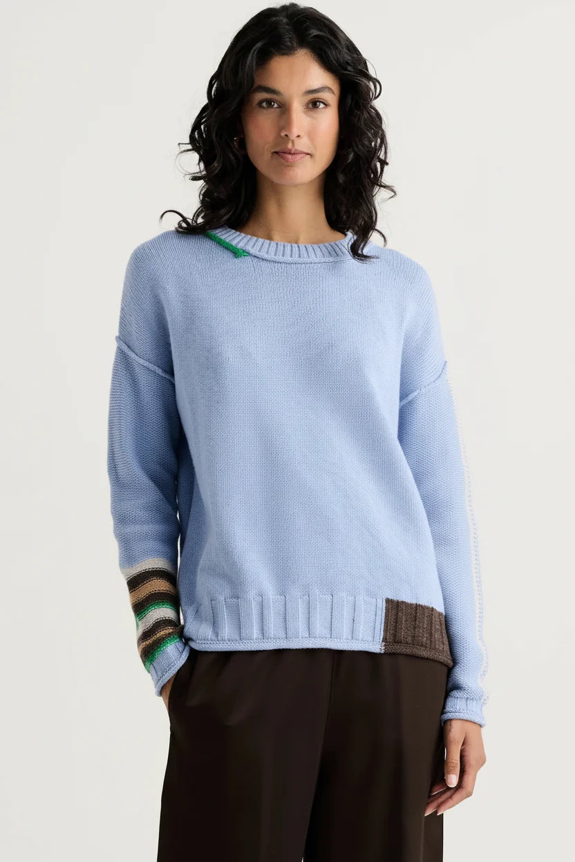 A woman modeling a light blue crewneck sweater with a ribbed hem, a brown patch pocket, and striped knit cuffs, paired with dark brown trousers against a light gray background.
