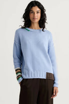 A woman modeling a light blue crewneck sweater with a ribbed hem, a brown patch pocket, and striped knit cuffs, paired with dark brown trousers against a light gray background.