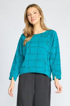 A woman modeling a teal long-sleeve blouse with a black geometric grid pattern, paired with dark gray wide-leg trousers, against a light gray studio background.