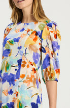 A woman wearing a short-sleeve blouse with a colorful watercolor floral print on a white base, featuring blue, purple, orange, yellow, and green tones.