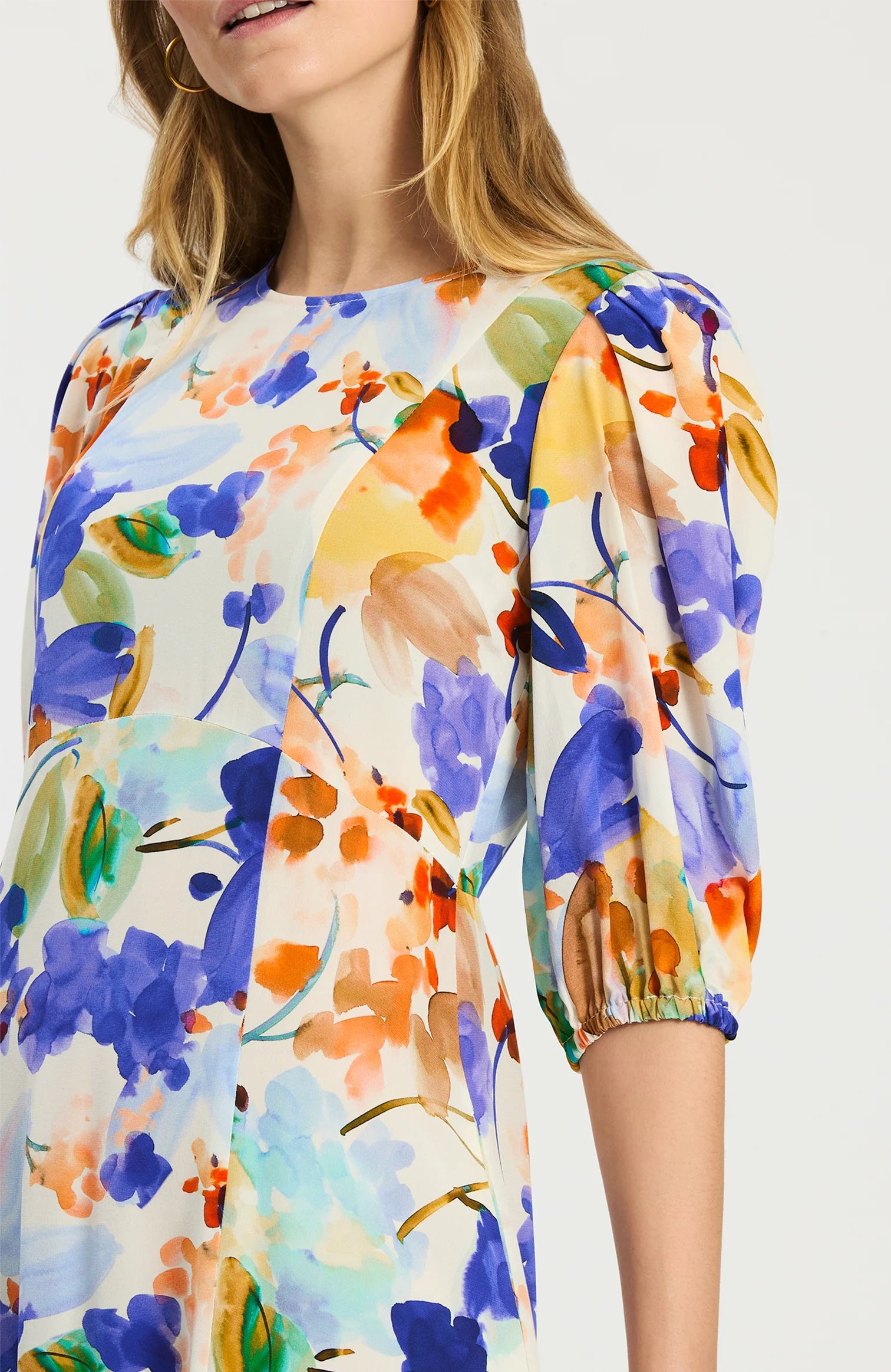 A woman wearing a short-sleeve blouse with a colorful watercolor floral print on a white base, featuring blue, purple, orange, yellow, and green tones.