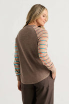 A woman shown from the back wearing a brown knit top with peach and blue striped raglan sleeves, paired with dark brown pants, against a light gray studio background.