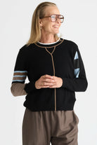 A woman wearing a black knit zip-front sweater with beige trim, striped sleeves, and brown pants, photographed against a light gray studio background.