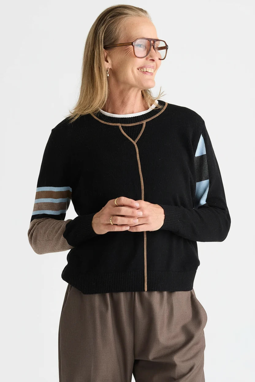 A woman wearing a black knit zip-front sweater with beige trim, striped sleeves, and brown pants, photographed against a light gray studio background.