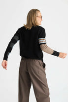 A model wearing a black knit sweater with color-blocked sleeves in gray and beige, paired with brown trousers, shown from a back-three-quarter angle against a light gray studio background.