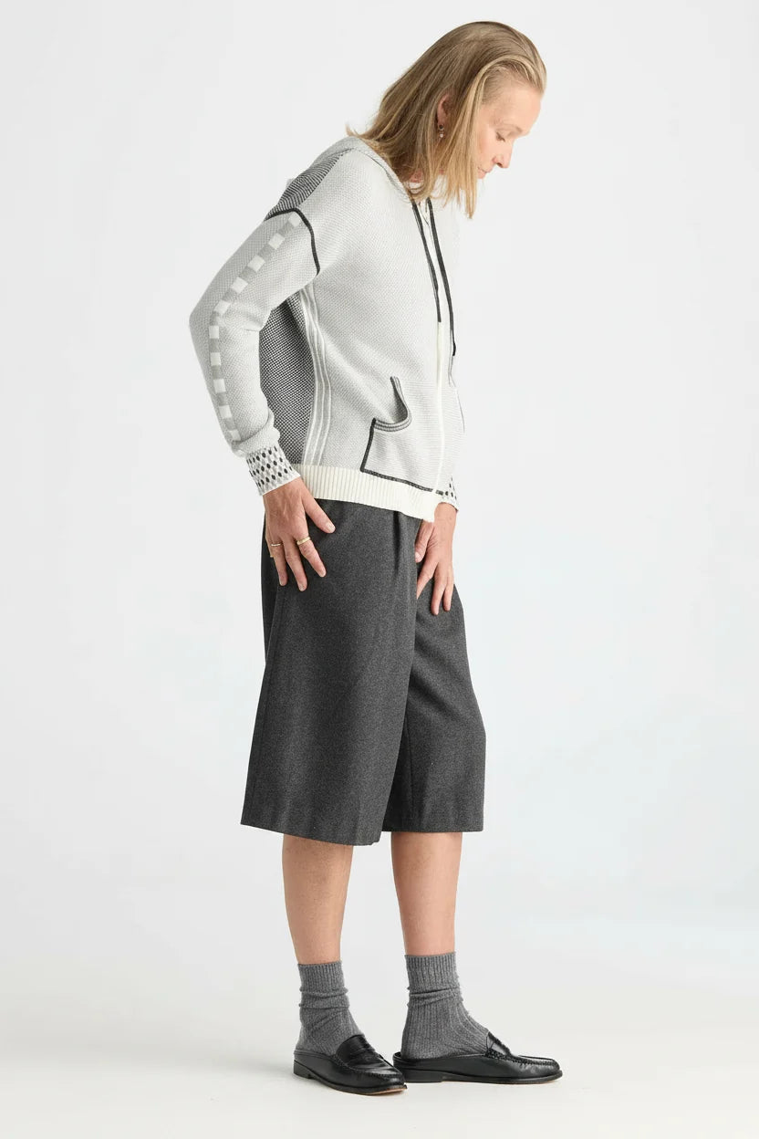 A model wearing a light gray and white knit zip-up hoodie with gray sleeve patches, dark gray knee-length shorts, gray socks, and black loafers, photographed against a plain light gray studio background.