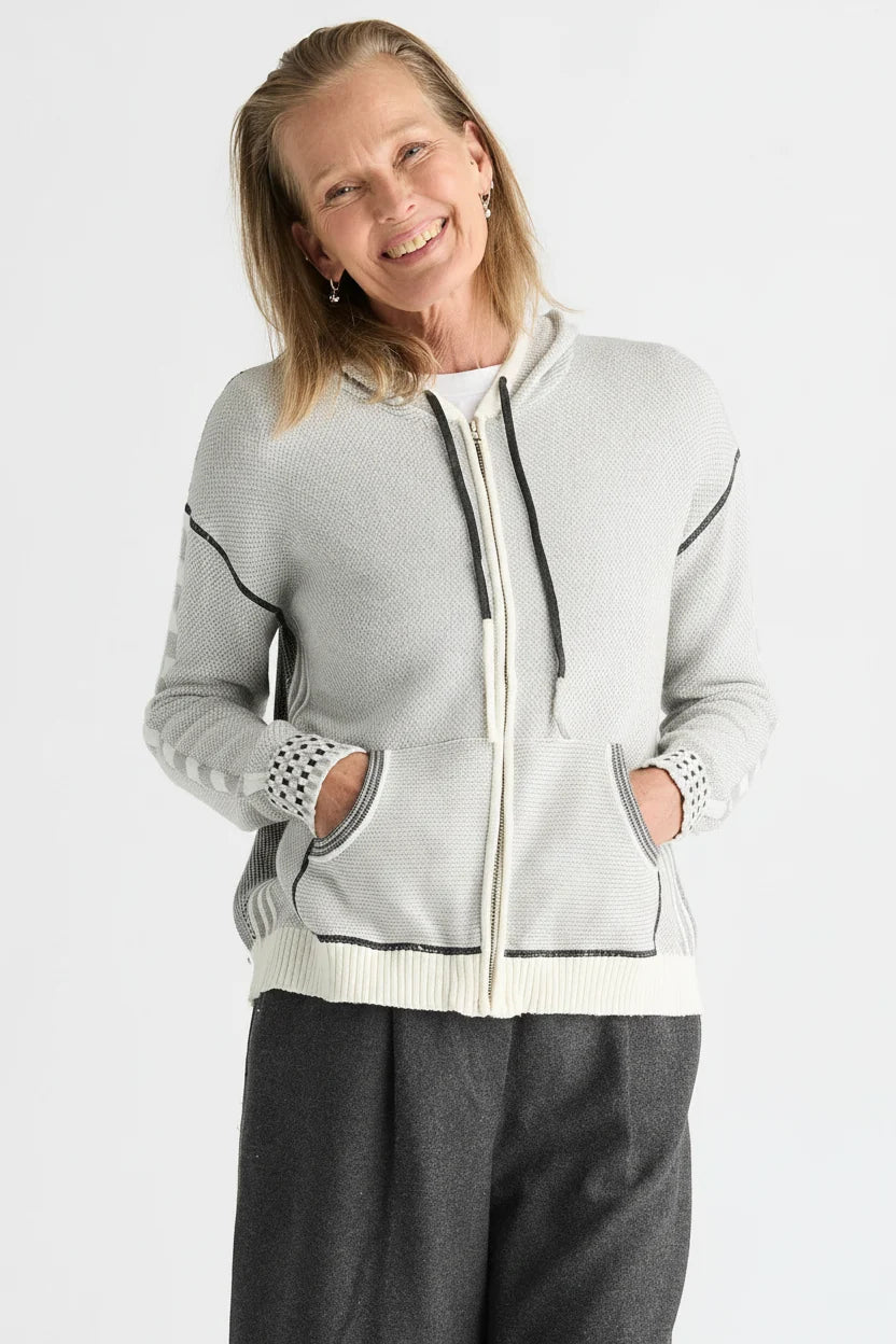 A woman wearing a light gray zip-up hoodie with black and white contrast stitching, black drawstrings, and white knit cuffs, paired with dark gray pants, against a plain white background.